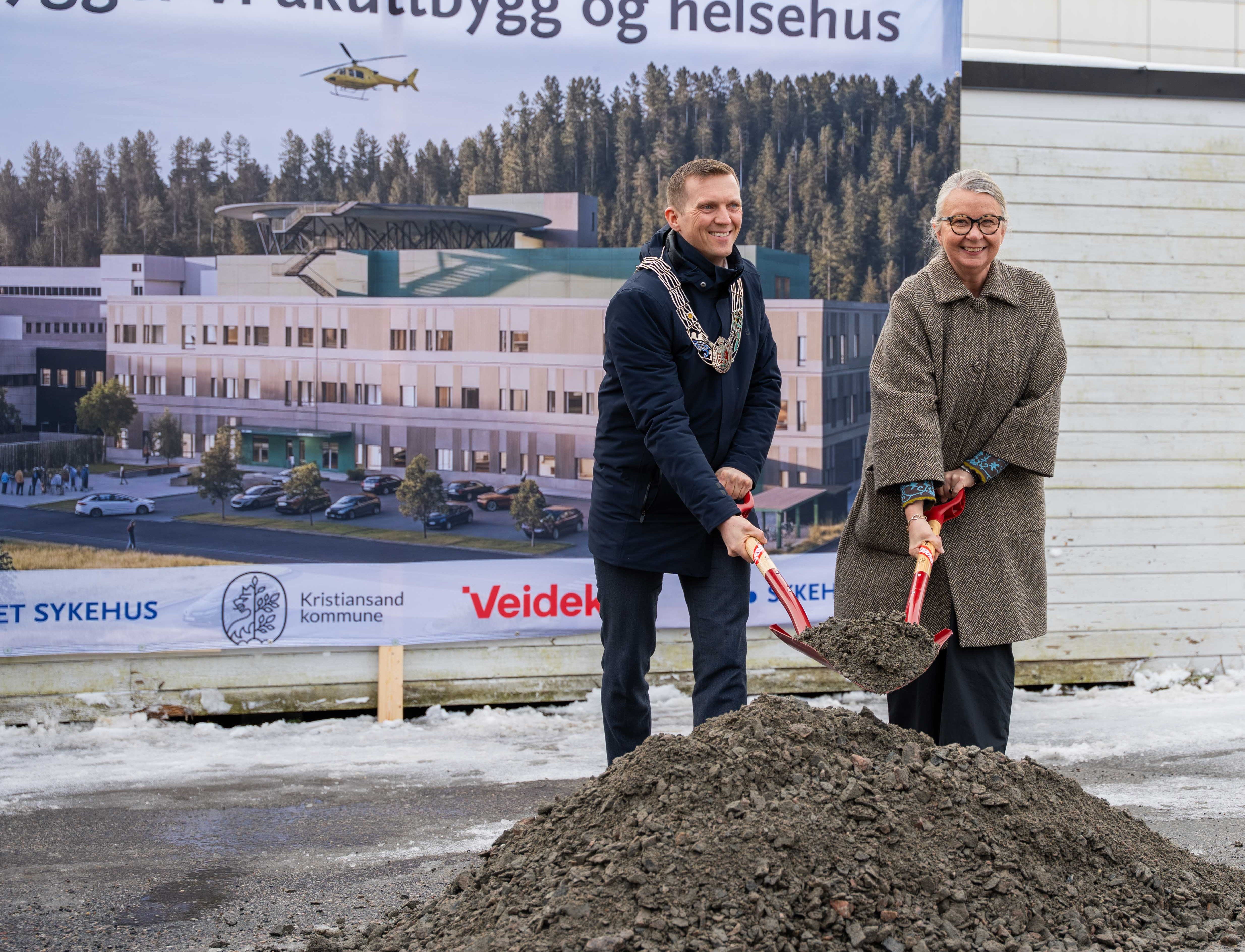 En mann og en kvinne som står ved en jordhaug med spade foran et stort banner med bilde av nytt akuttbygg