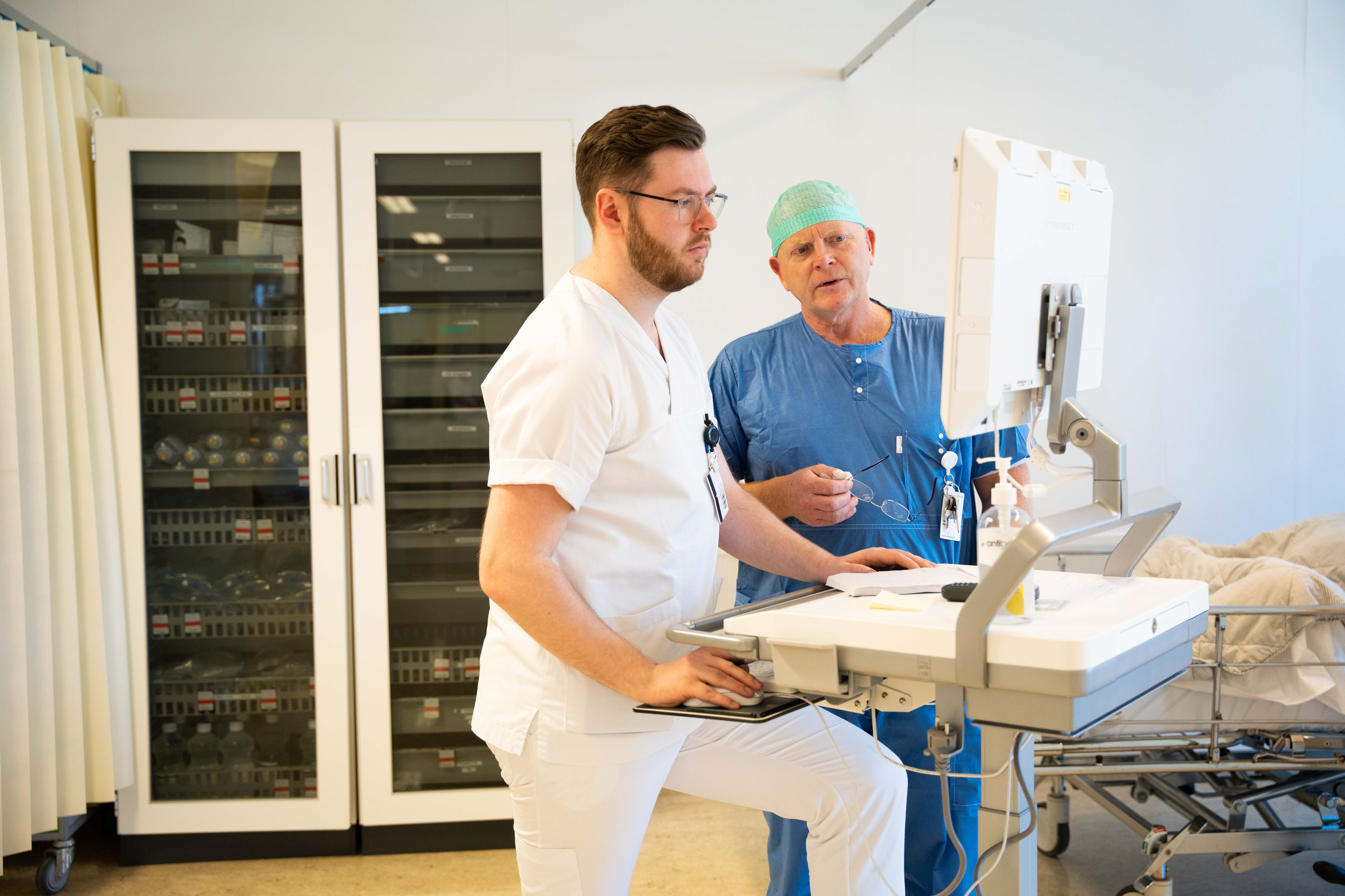 To menn kledd i hvitt og blått samtaler sammen inne på intenisv.