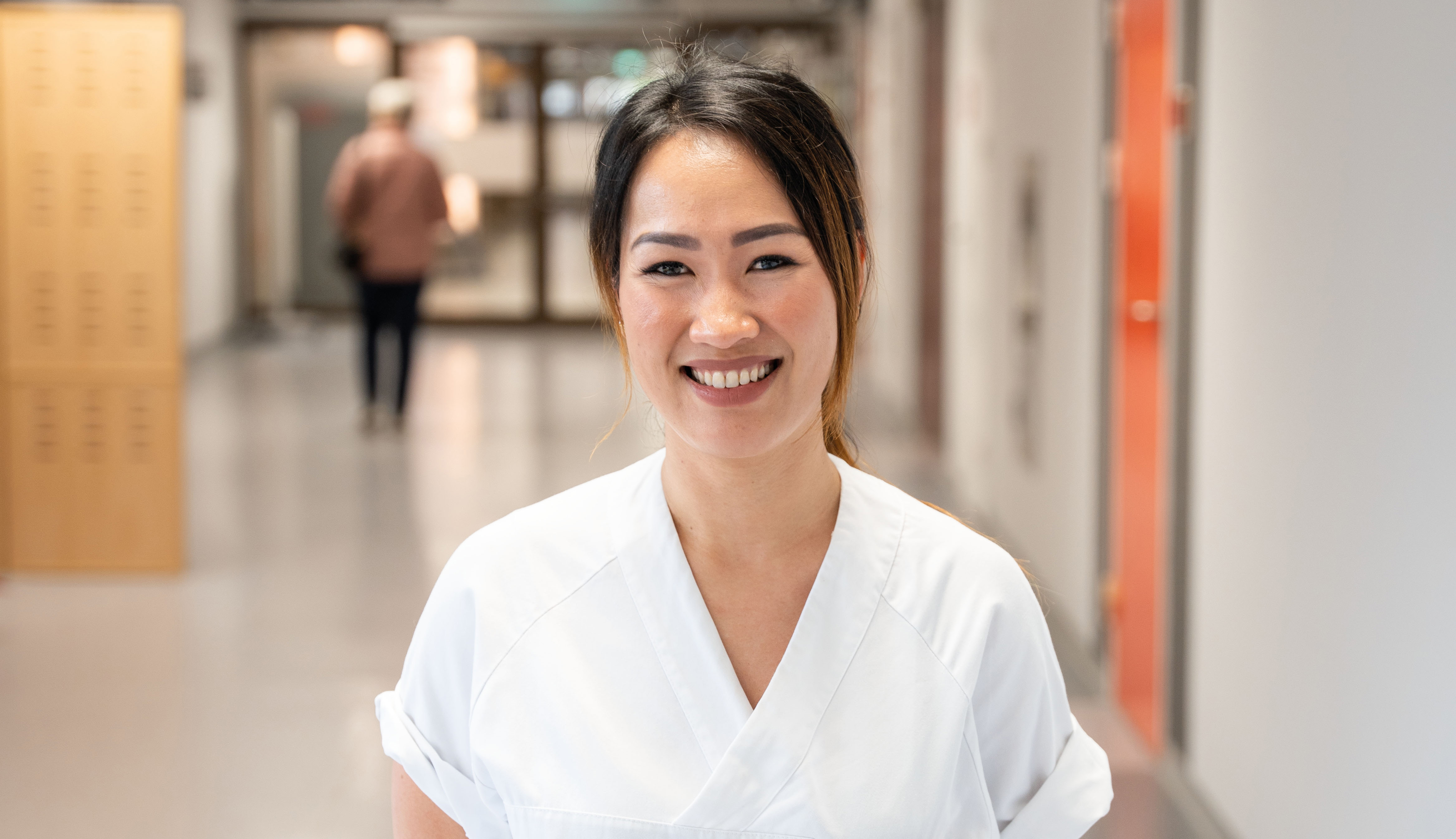 Sykepleier kledd i hvit uniform står smilende i en korridor på sykehuset.