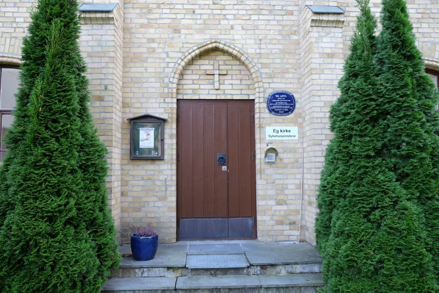 Kirkedør omkranset av grønn trør. To trappetrinn opp til døren. Foto.