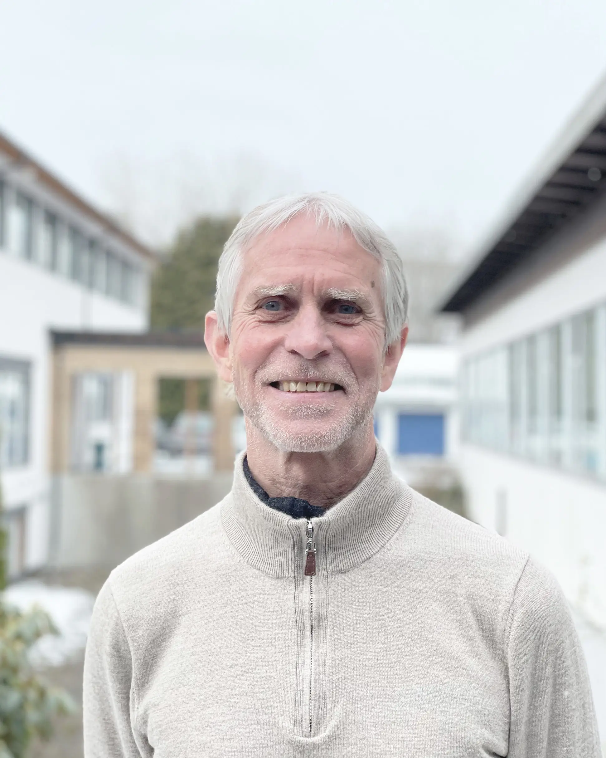 Mann står utendørs i hvit genser og smiler mot kamera. Foto.