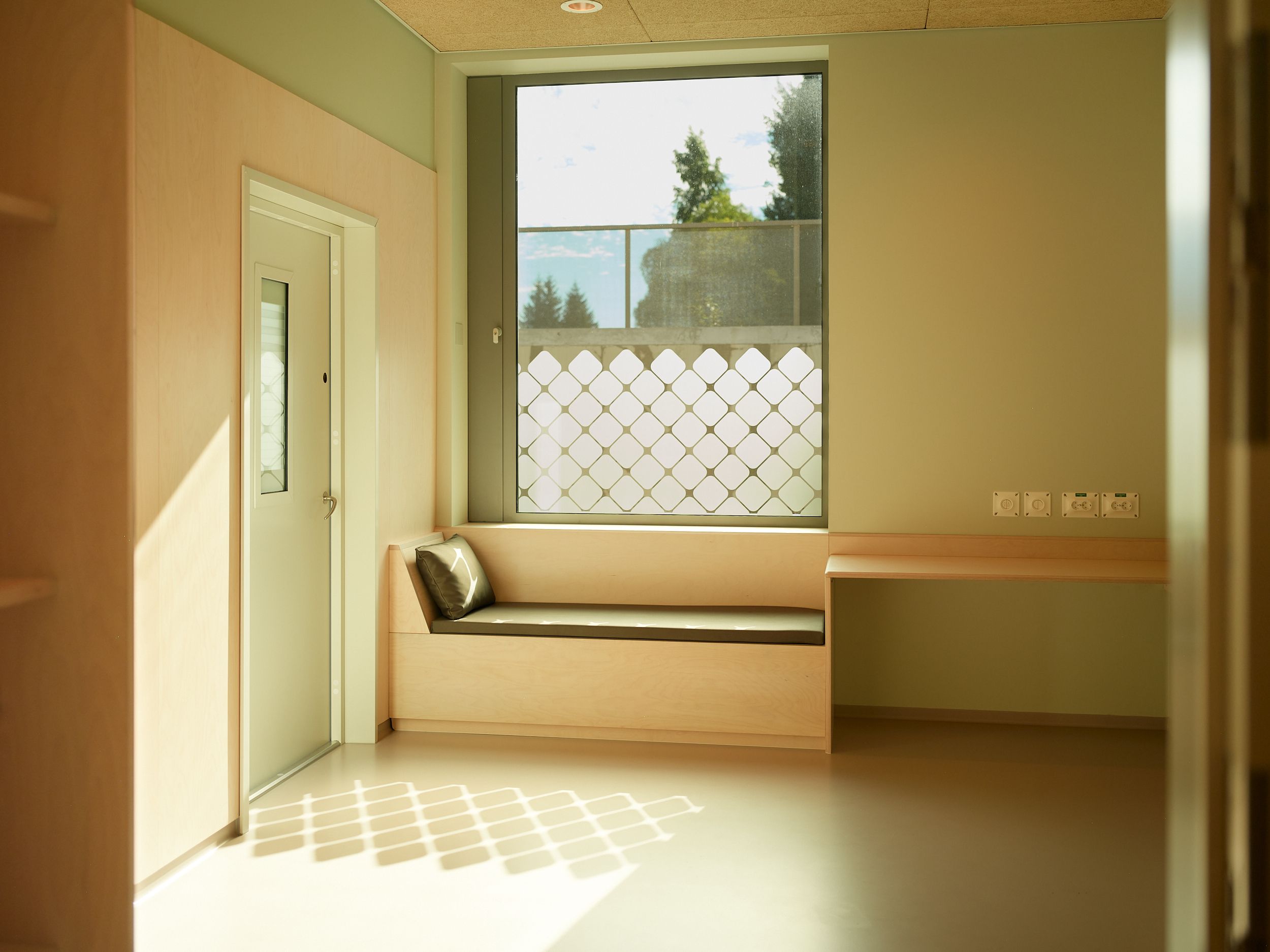 Pasientrom med stort vindu, daybed og skrivebord. Sollyset reflekteres i rommet. Foto.